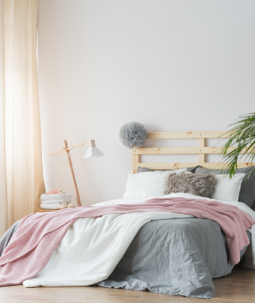 Habitación con cama de matrimonio con cubrecolchón en color gris y mantita en rosa, palmera y lámpara en la mesa de noche, cabecera de madera natural