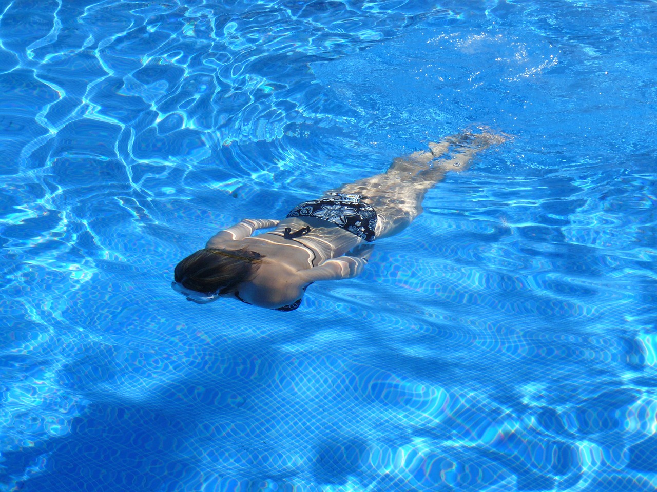 Esta refrescante foto captura a una mujer en bikini nadando con gracia en una piscina. El agua cristalina resalta su figura mientras se desliza suavemente a través de ella.